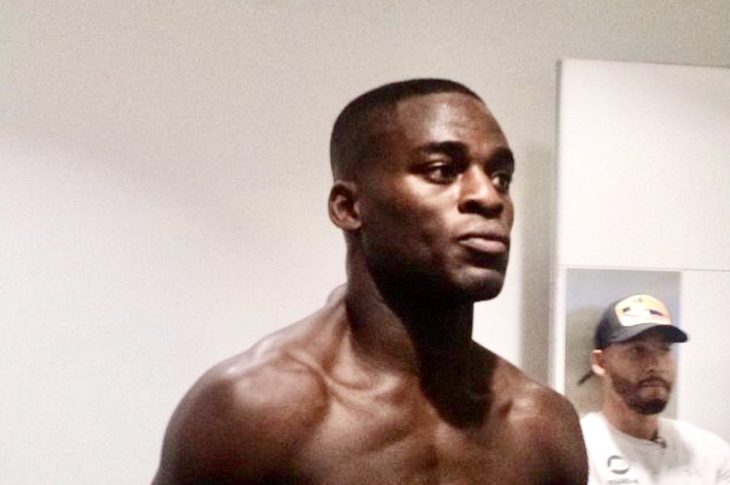 Boxer Joshua Buatsi readies himself in his dressing room. He is topless, and is wearing black shorts with a red waistband and black boxing gloves.