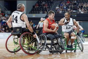 Wheelchair basketball athlete James Rose (C) in a red Great Britain jersey holds the ball, marked by two New Zealand defenders (L and R) in white jerseys