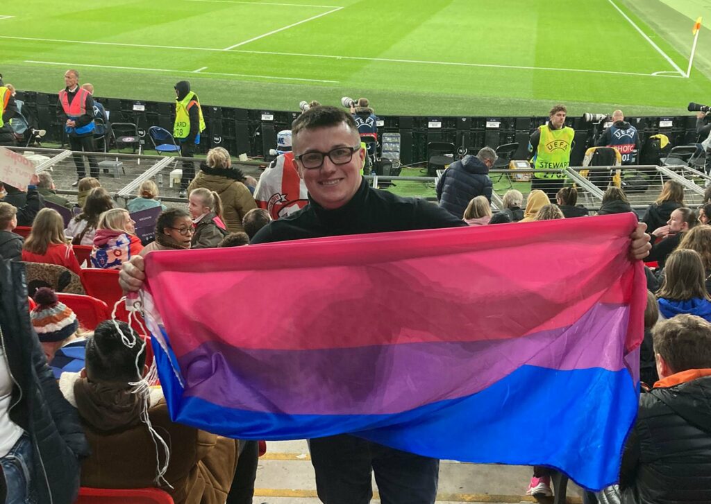 Michael McCann holds up the bisexual flag while in the crowd of a football stadium