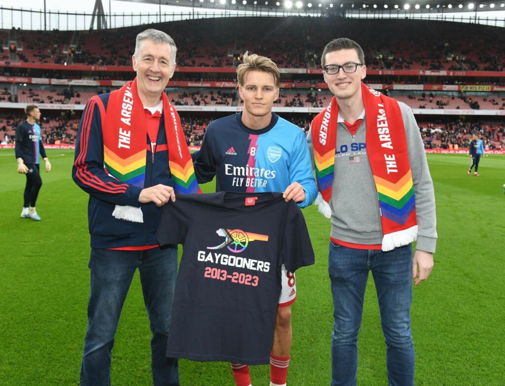 Arsenal captain Martin Odegaard with Carl Fearn and Jacob Jefferson of GayGooners on the Emirates pitch holding a GayGooners t-shirt making Arsenal a safe space
