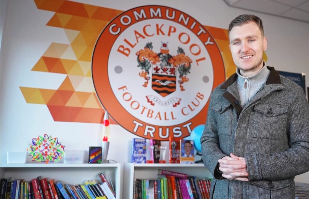 Nick Ransom stands in front of a wall mural showing Blackpool City Community Trust badge, he often presents on neurodivergence in the sports media.