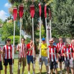 Twelve crew members, all wearing Brentford shirts, stand in a back garden with their oars pointed to the sky.