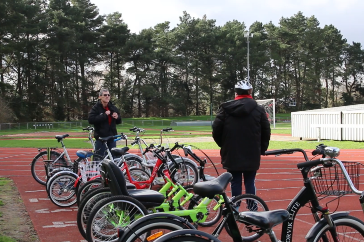 A row of tricycles are lined up on a red athletics track with a man wearing a bike helmet and Wheels for All volunteer stood next to them.