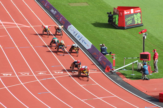 Wheelchair racers approach cross the finish line on red athletics track.